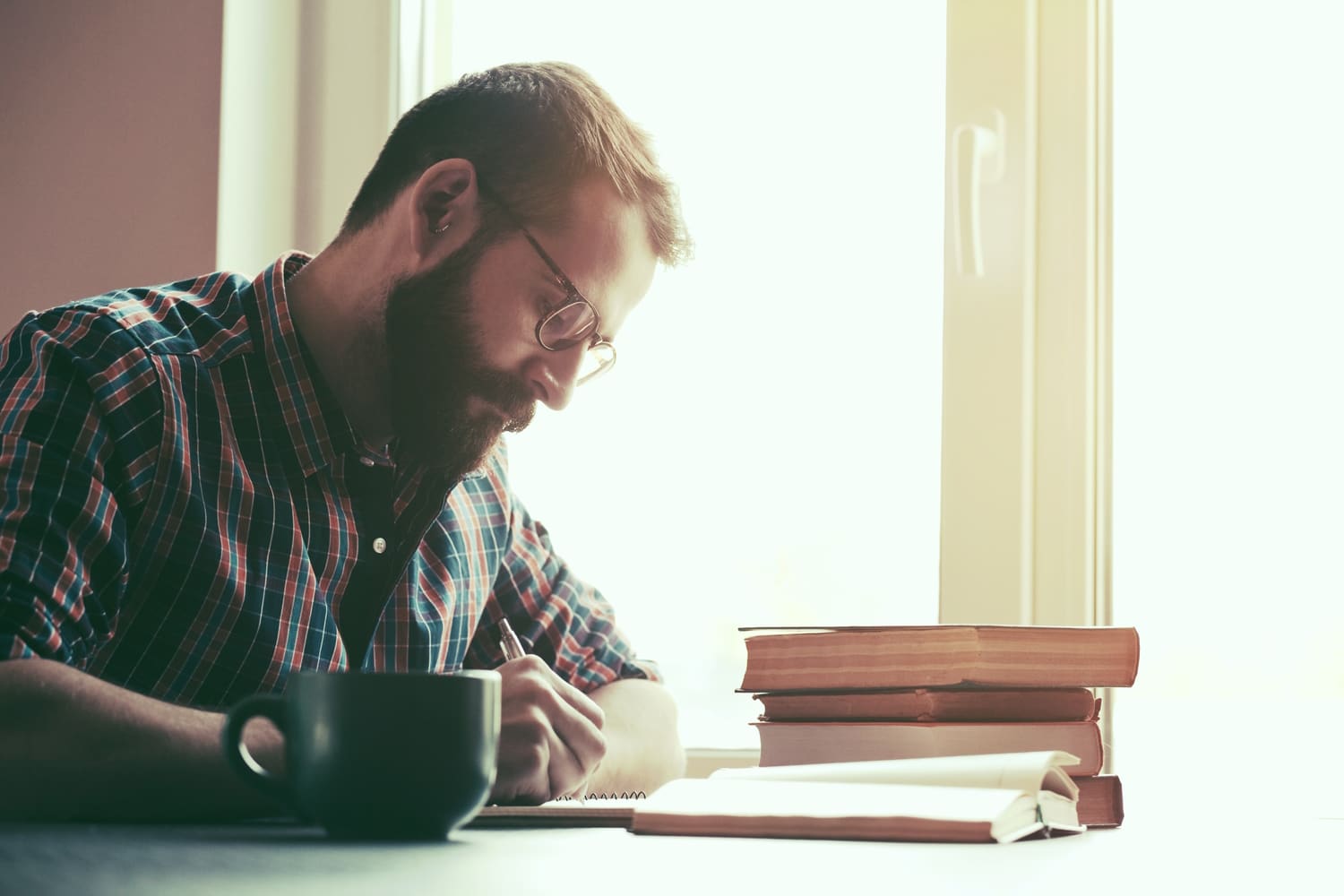 Bearded man writing with pen and reading books at table