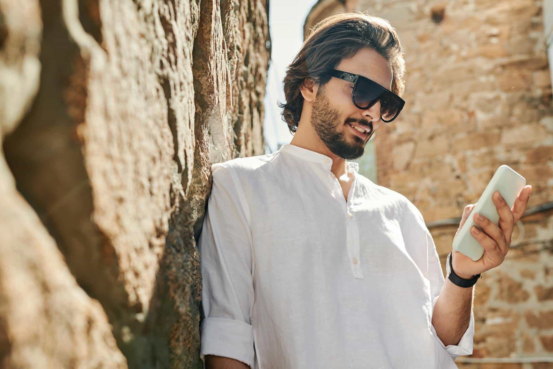 A Bearded Man in White Long Sleeves Wearing Sunglasses Using His Cellphone while Leaning on a Stone Wall.