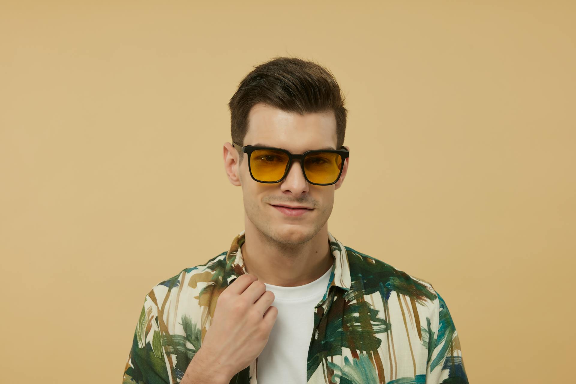 A man wearing orange tinted sunglasses holding the collar of his floral printed shirt.