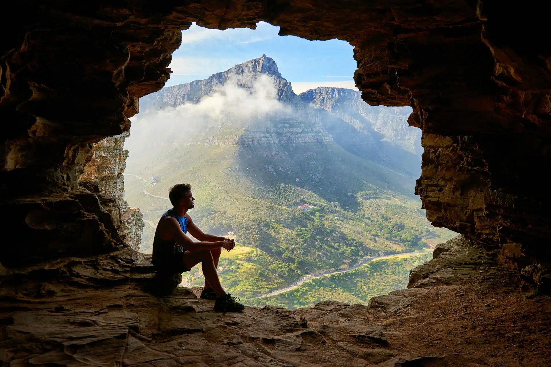 Man sitting at the entrance of a cave with mountains and a valley in the background.