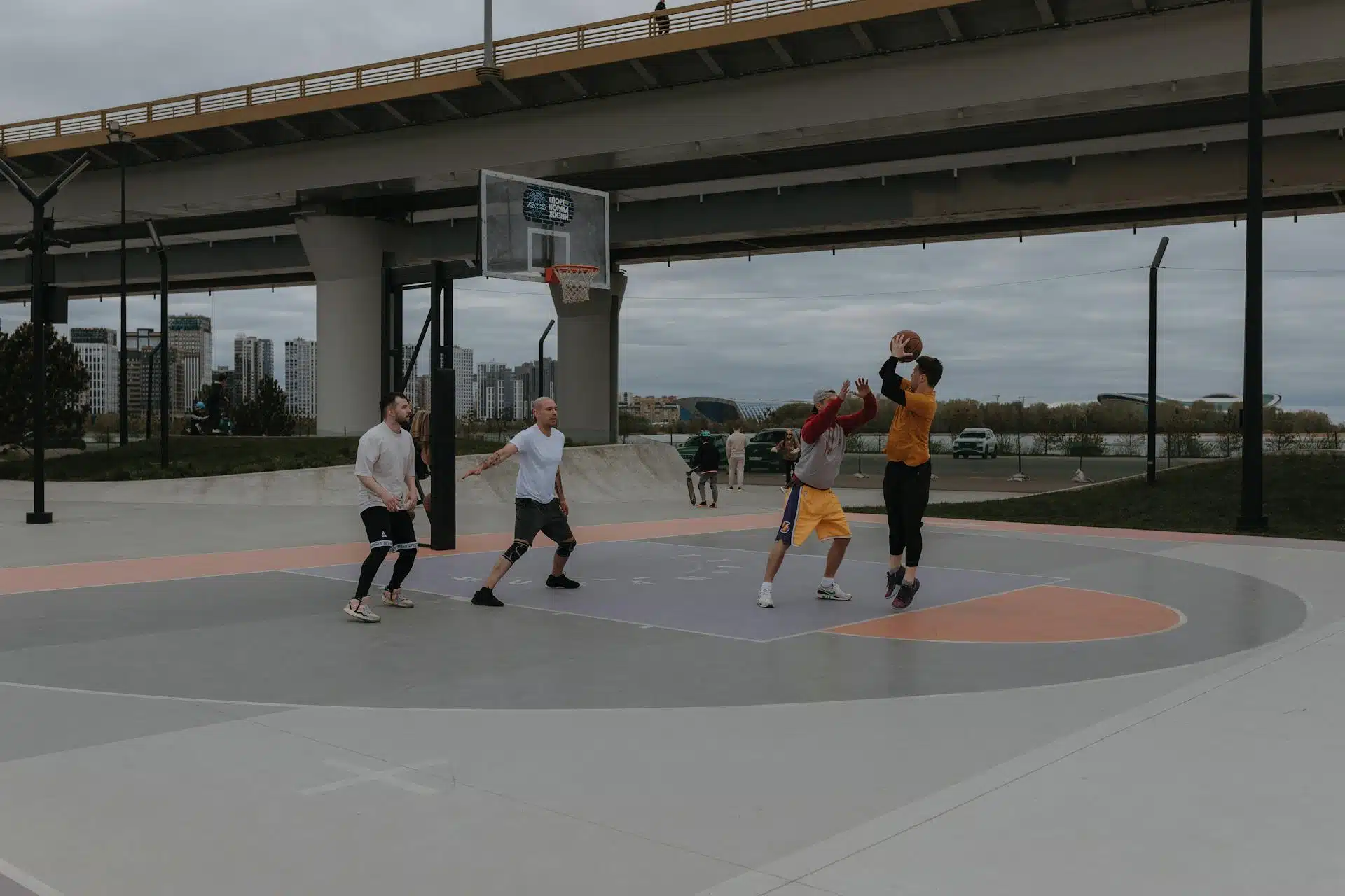 A group of four friends playing basketball at a court by a bridge.