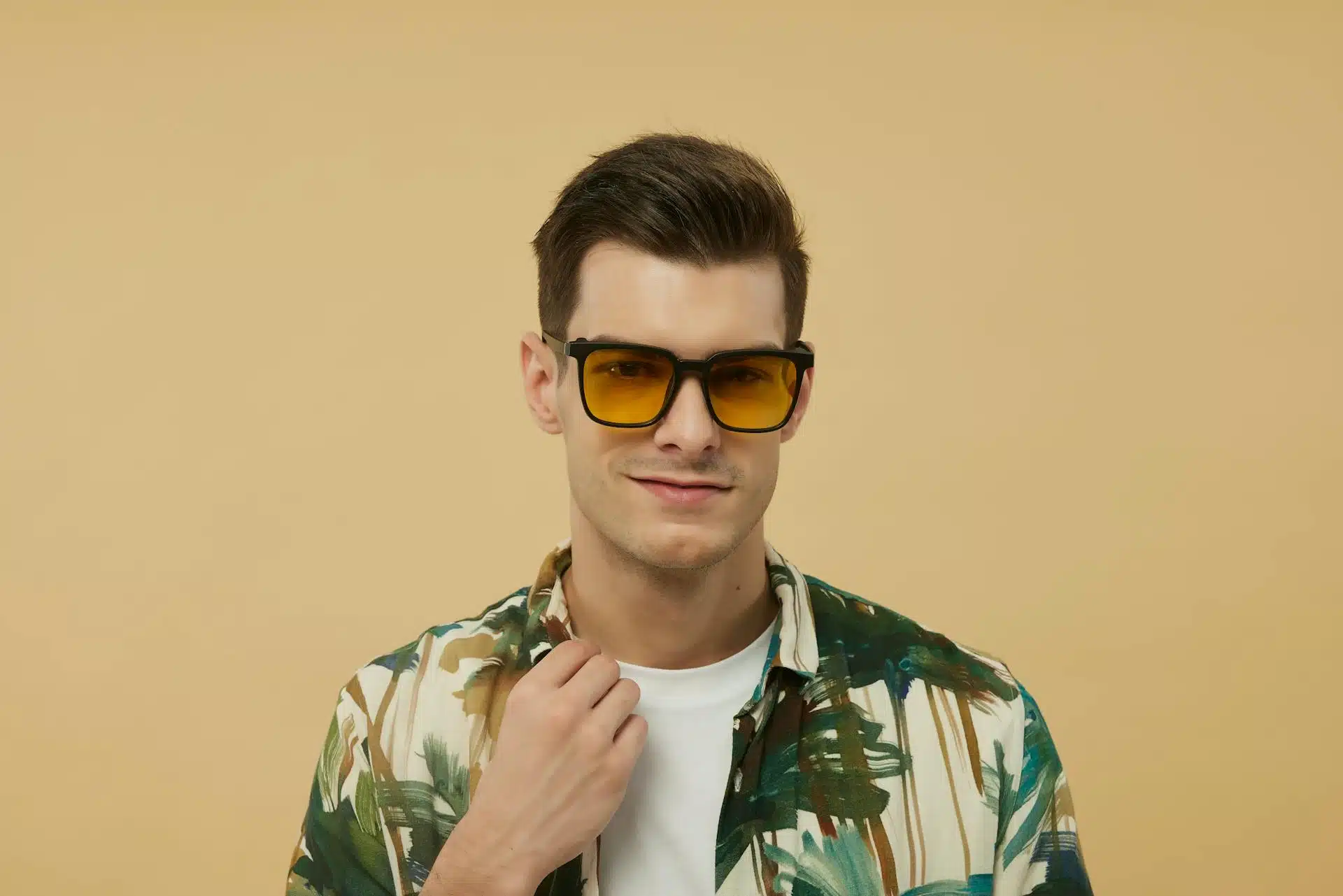 A man wearing orange tinted sunglasses holding the collar of his floral printed shirt.