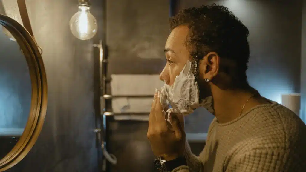 A curly-haired man applying shaving foam to his beard while standing in front of a round mirror.
