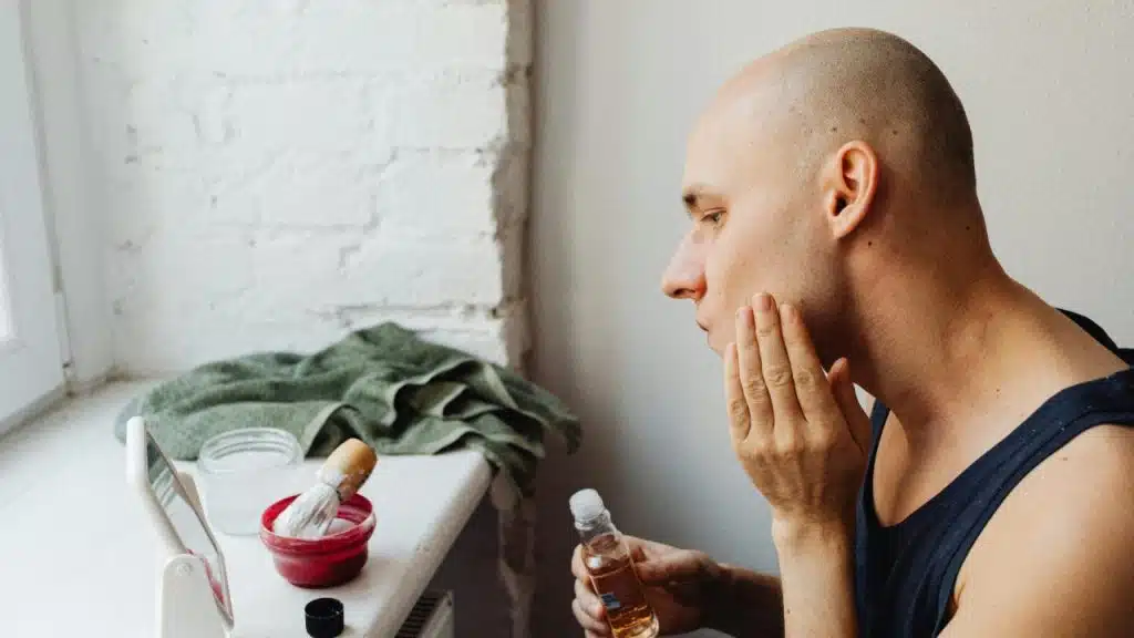 Bald man in a black tank top applying aftershave with a square-shaped mirror, a shaving brush and a towel placed on the shelf in front of him.