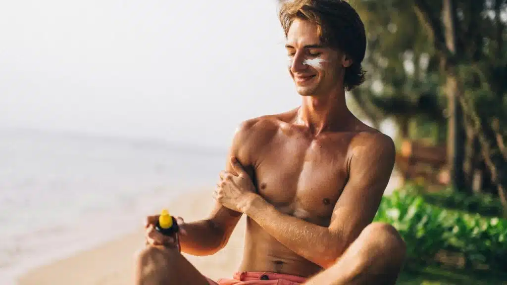 Shirtless man in shorts sitting on the beach while applying sunscreen to his arm.