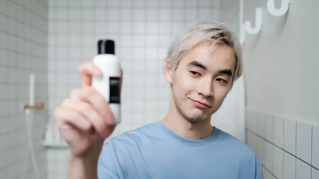 Man with dyed hair standing in a bathroom, holding up a bottle and looking at it.