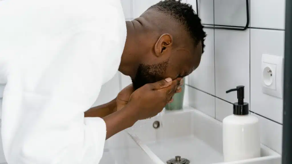Man in a white bathrobe washing his face over a bathroom wash basin.