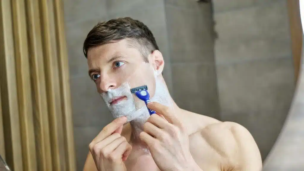 Shirtless man with a toned physique shaving his face using a blue razor.