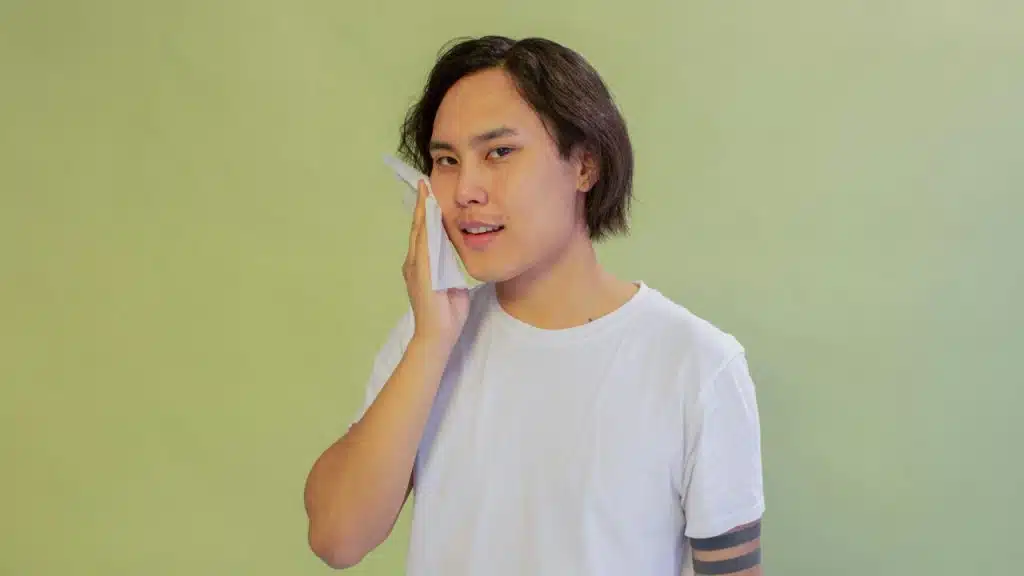 Man in a white T-shirt wiping his face with a tissue against a green background.