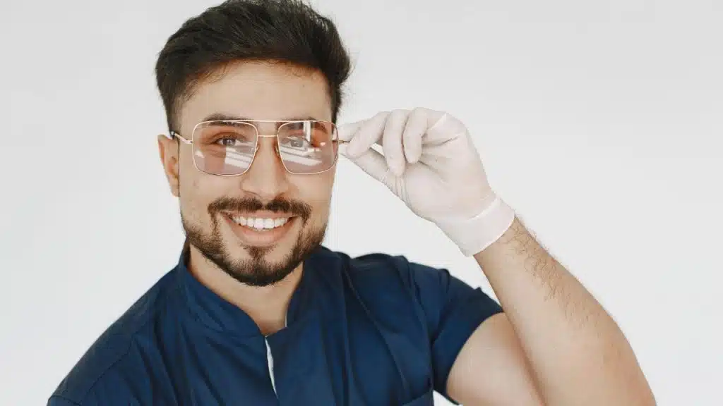 Smiling man in blue scrubs adjusting his sunglasses with a gloved hand.