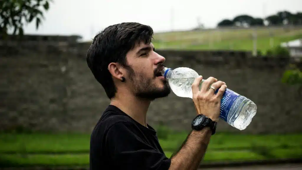 Man in a black t-shirt with a watch on his right wrist drinking water from a plastic bottle with his eyes closed.