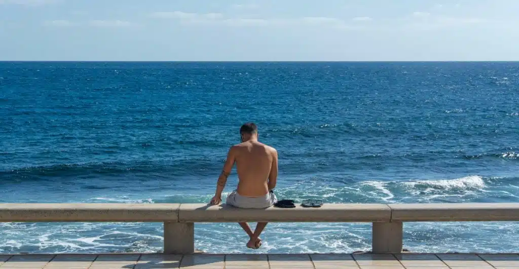 Shirtless man sitting on a concrete railing by the ocean, with sunglasses and clothes placed beside him.