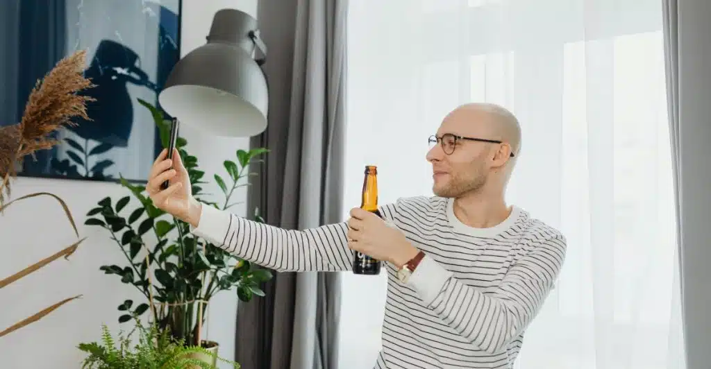 Bald man in a striped shirt taking a selfie while holding a bottle of beer.