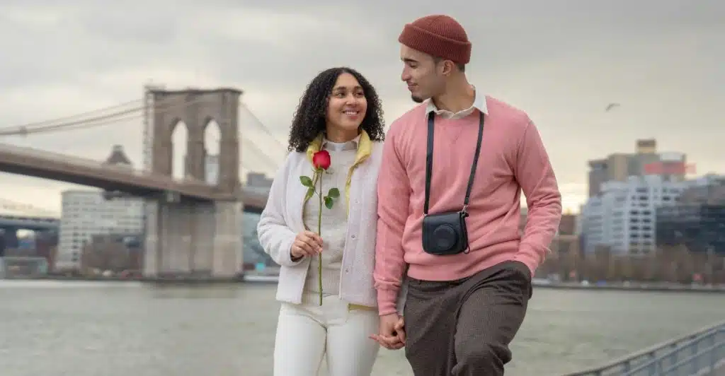 Romantic couple holding hands by a river, with a bridge and city buildings in the background.