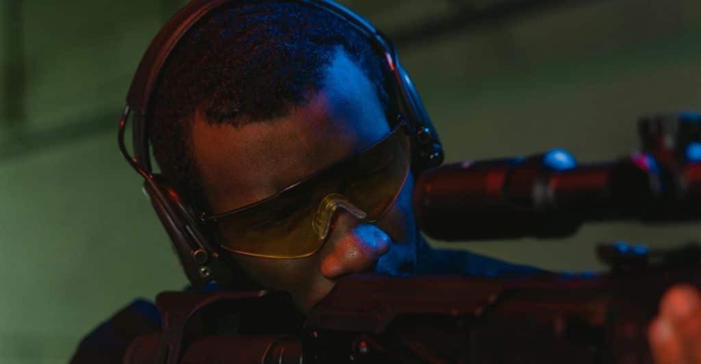 Man in tactical gear aiming through the scope of an assault rifle.