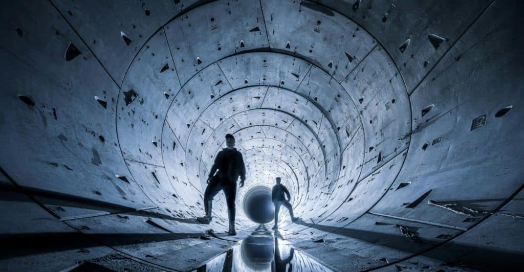 Silhouettes of two men standing in a futuristic tunnel with reflective water on the ground.