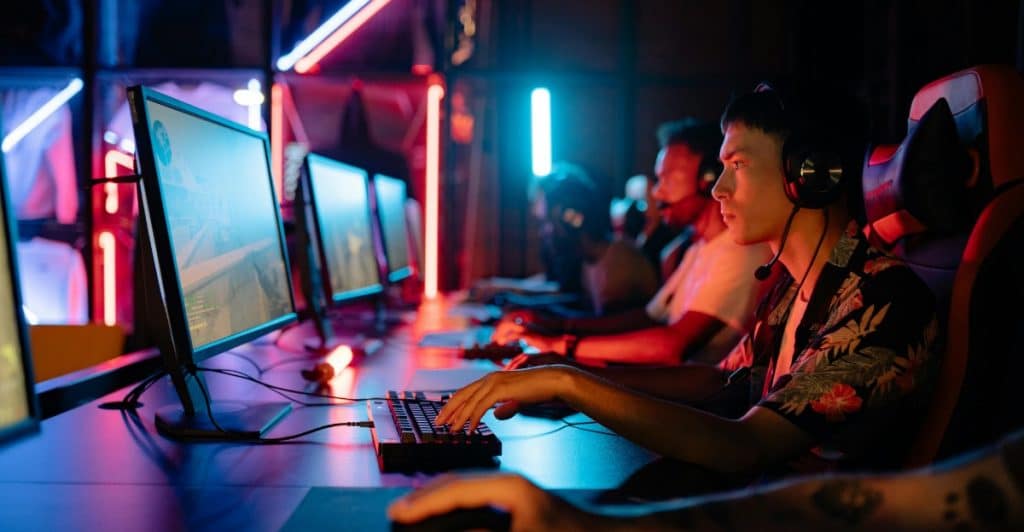 Focused gamers wearing headsets playing on desktop computers in a dimly lit gaming arena with neon lights.