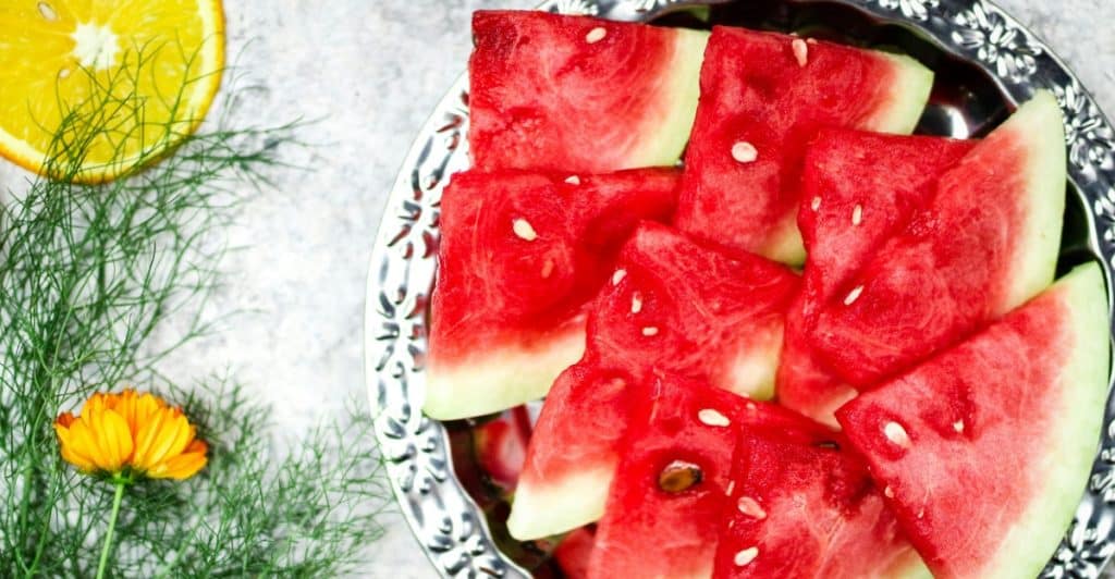 Plate full of watermelon slices placed beside yellow flowers and a slice of orange.
