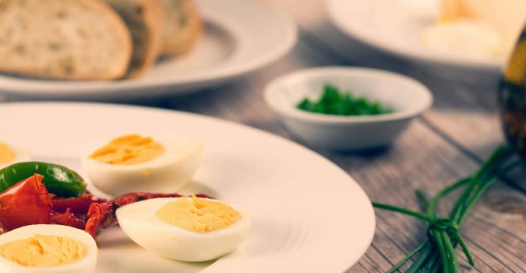 Two sliced hard boiled eggs with vegetables on a plate with a plate full of sliced garlic bread in the background.