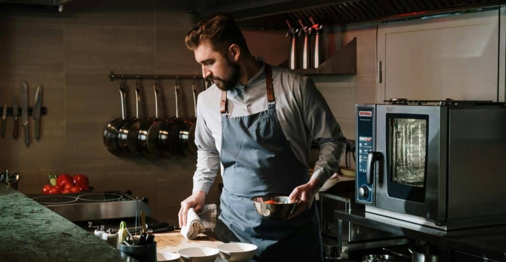 Male chef in a professional kitchen preparing food while holding a metal mixing bowl and towel.