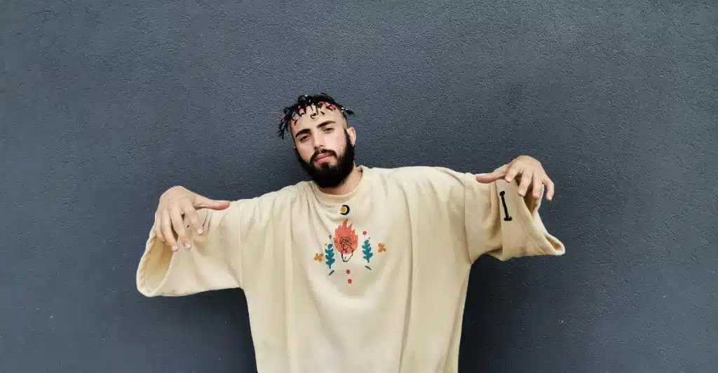 A man with a beard and braided hair, wearing an oversized tan t-shirt with a graphic design, standing against a grey wall.