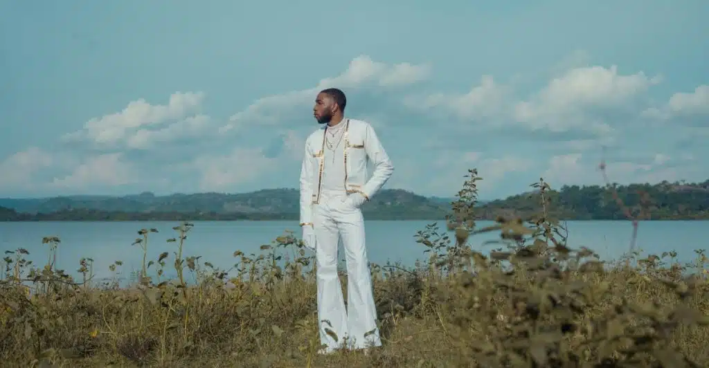 A man wearing a white outfit with a jacket and wide-leg pants, standing outdoors by a lake or large body of water.
