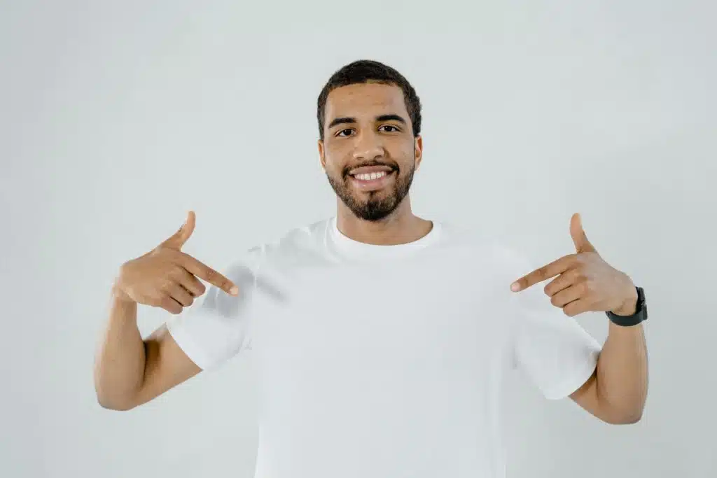 A smiling man in a white t-shirt and dark pants, pointing upwards with both hands against a white background.