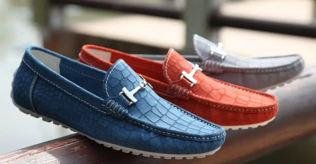 Three leather loafers in blue, orange, and grey with metal buckles, lined up on a wooden surface.