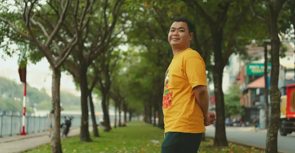 A man in a yellow graphic t-shirt, smiling and looking over his shoulder, standing in an tree-lined outdoor area.