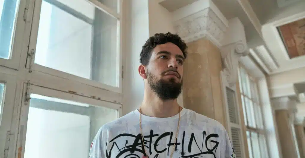 Close-up of a man wearing a white t-shirt with large black and pink graphic logos, looking away from the camera.