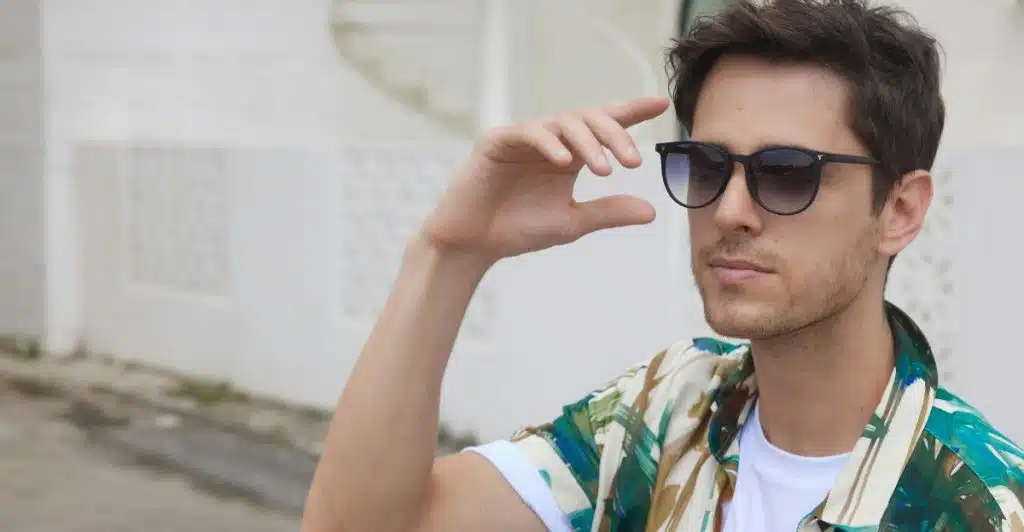 A man wearing a green and blue abstract pattern short-sleeve shirt over a white t-shirt, and dark shorts, sitting outdoors while adjusting his sunglasses.