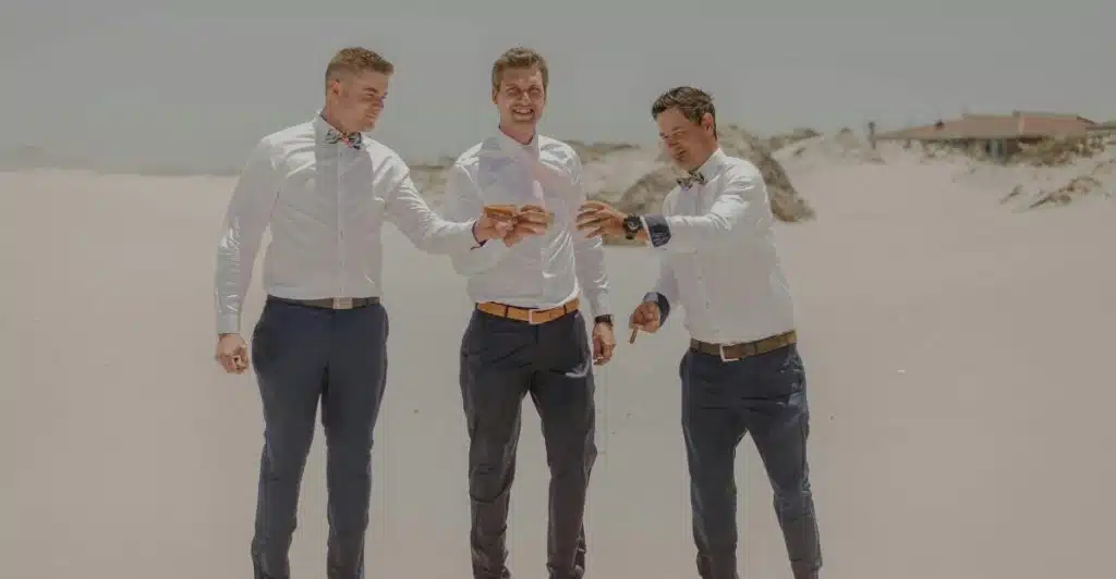 Three men in white long-sleeve shirts and dark trousers standing on a sandy beach.