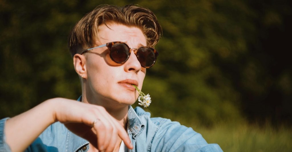 Close-up of a man in a light blue open shirt over a white tank top, wearing sunglasses and a necklace, with a flower in his mouth.