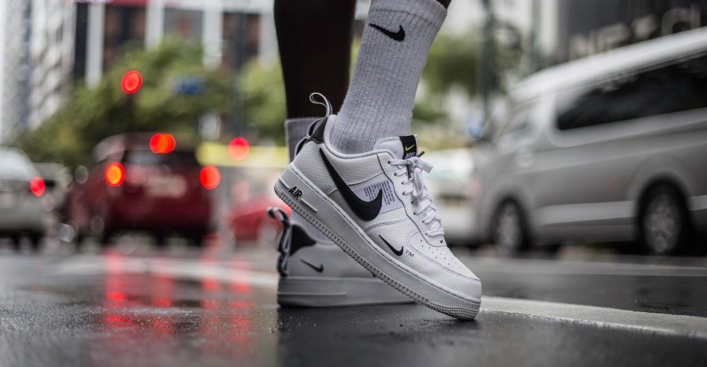 Close-up of feet wearing white sneakers and grey socks with a black Nike logo, standing on a wet street.