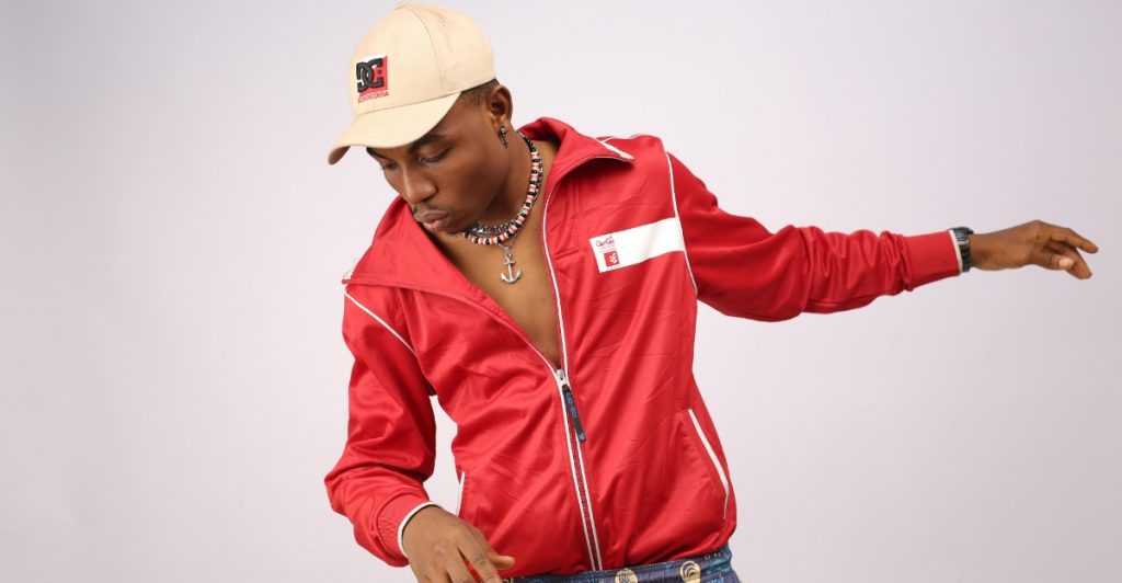A man wearing a red zip-up track jacket over a light shirt, patterned shorts, a necklace, and a baseball cap, posing with his arms out.