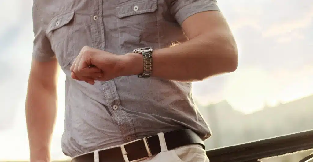 Close-up of a person wearing a grey short-sleeved shirt and white pants, looking at their wristwatch while standing outdoors.