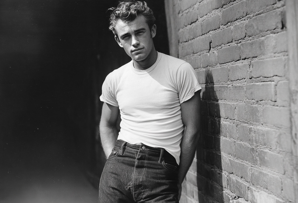 Vintage photo of a James Dean in a white T-shirt and jeans, half-tucked, leaning against a brick wall with effortless cool.