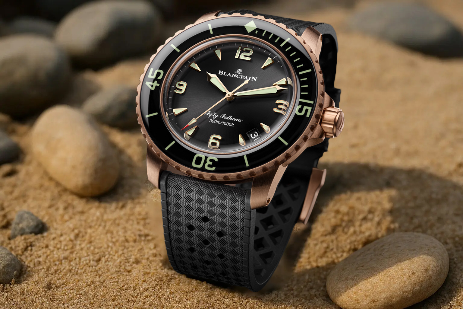 Blancpain Fifty Fathoms dive watch with a rose gold case and black dial, photographed on sand with smooth river stones.