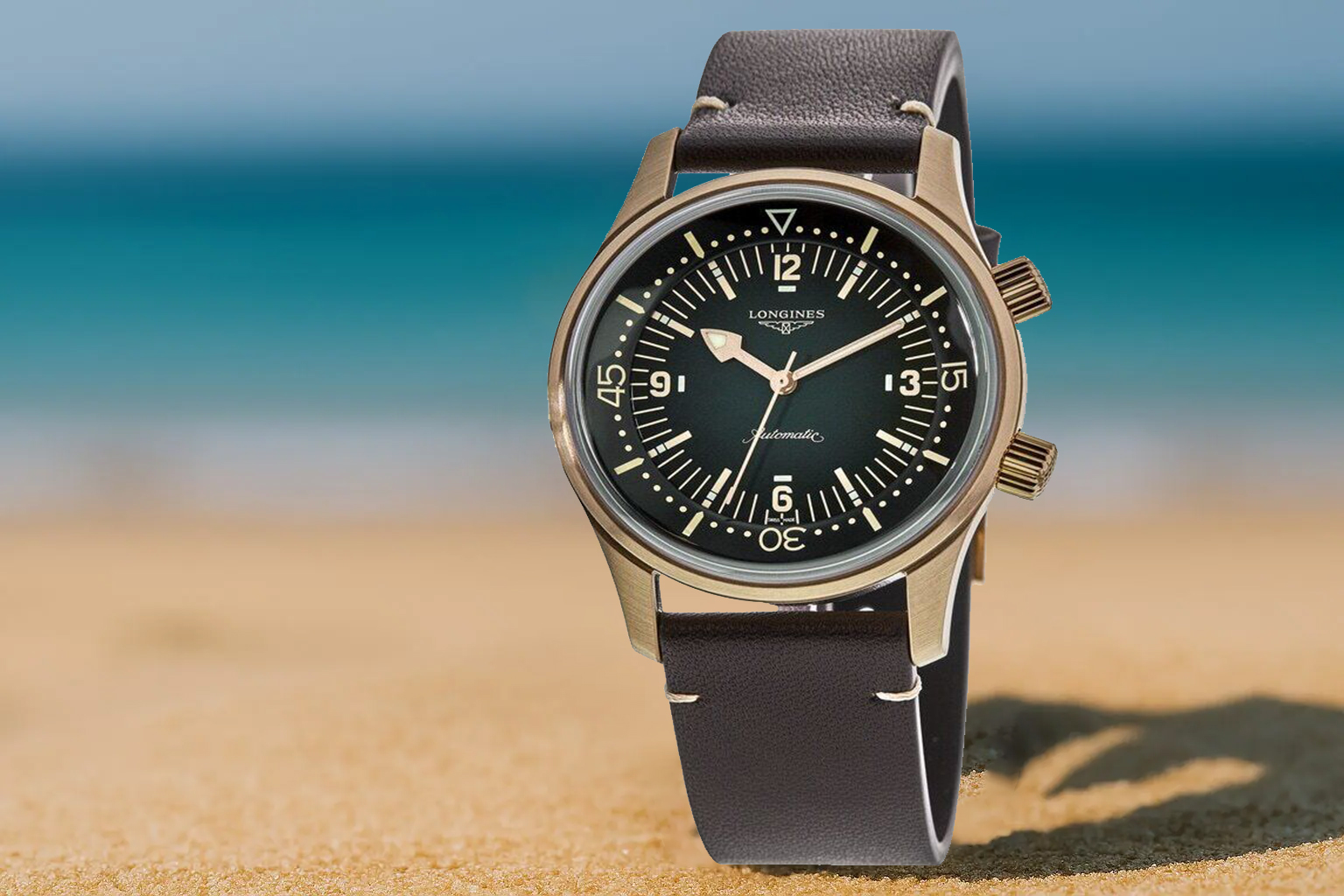 Longines Legend Diver watch with a bronze case and black dial, positioned upright on a sandy beach with turquoise sea in the background