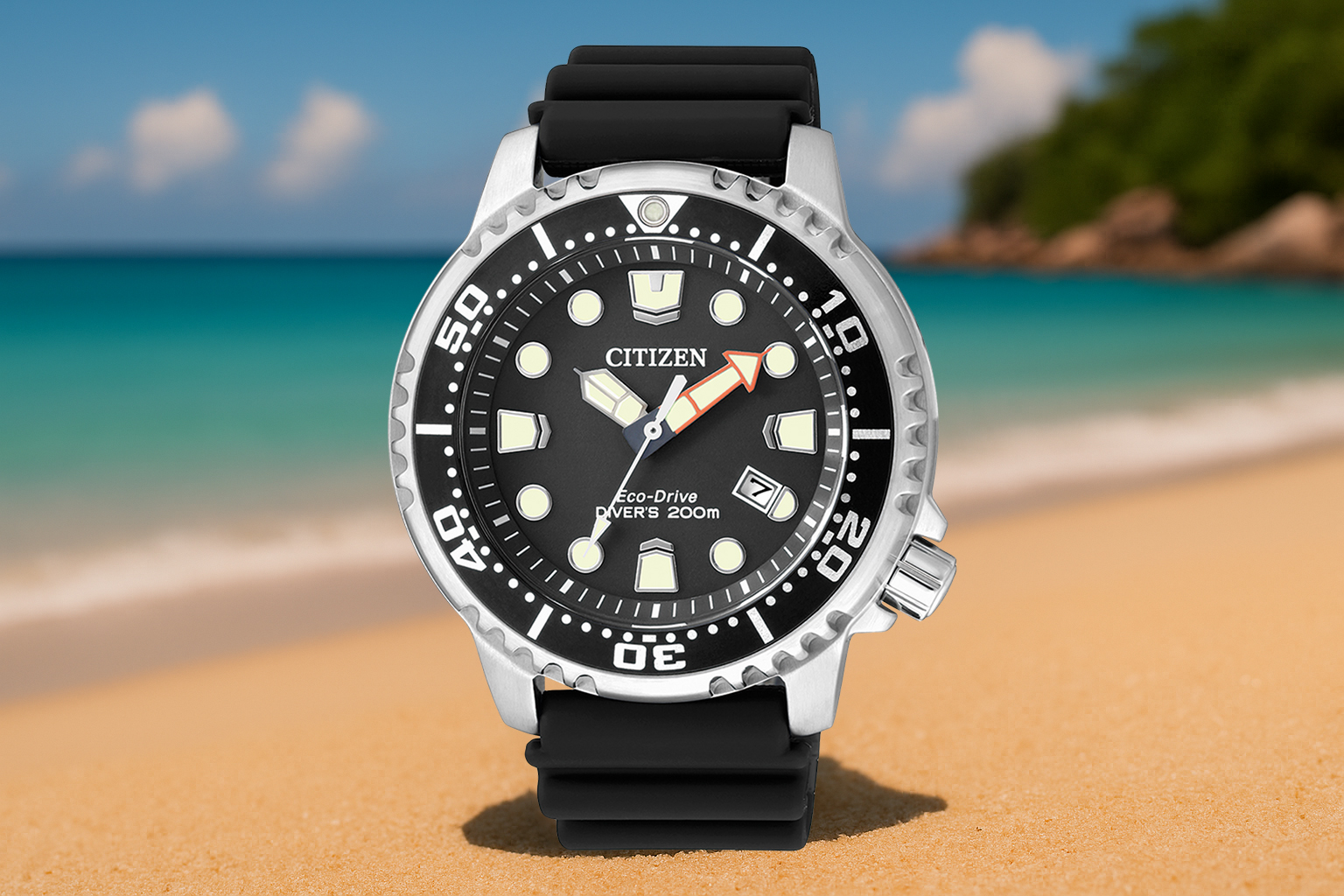 Citizen Promaster Diver Eco-Drive watch with a black dial and rubber strap, standing upright on golden sand with turquoise water and a tropical coastline in the background