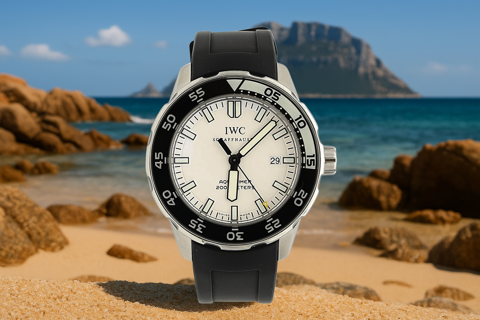 IWC Aquatimer 200 dive watch with a white dial and black bezel, displayed on sandy terrain with rocky shores and a coastal mountain in the background