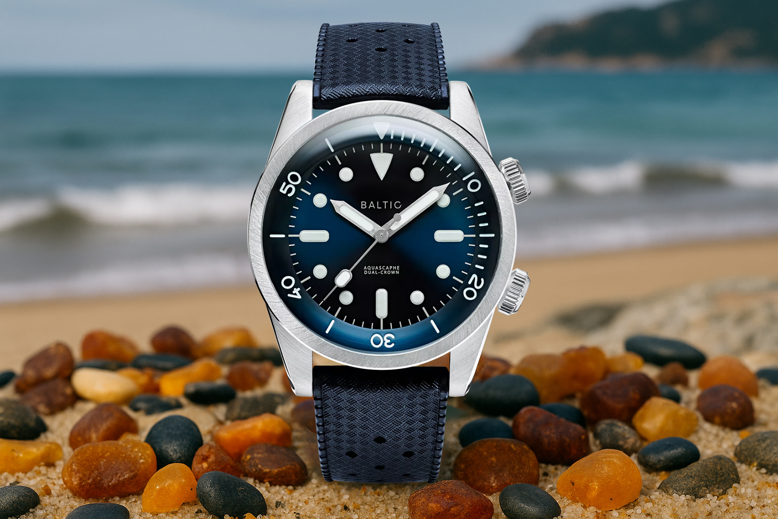 Baltic Aquascaphe Dual Crown dive watch with a gradient blue dial and navy strap, placed on Baltic sand surrounded by amber and black pebbles with waves in the background