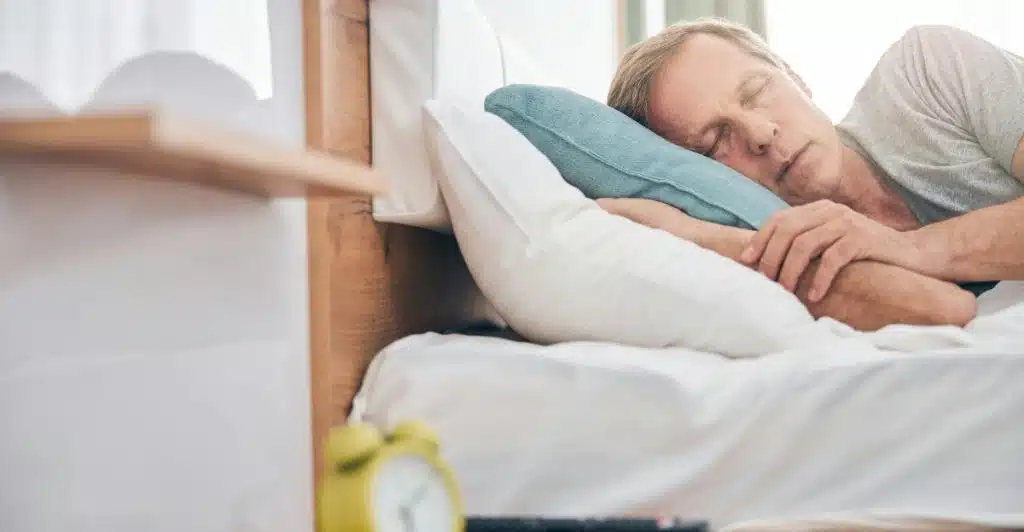 A person is sleeping peacefully in a bed with a yellow alarm clock, a phone, and a remote control on the bedside table.