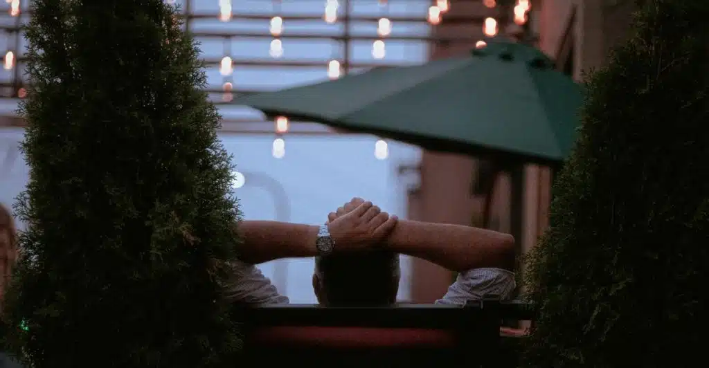 A person sitting on an outdoor bench with their arms behind their head, appearing relaxed.