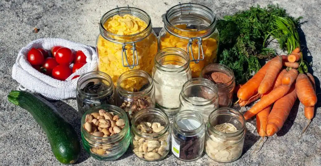 Various foods in glass jars arranged with fresh vegetables on a stone surface.