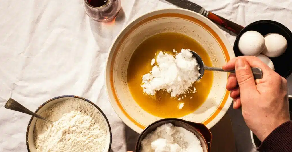 A person is adding a spoonful of white sugar to a bowl of other ingredients.