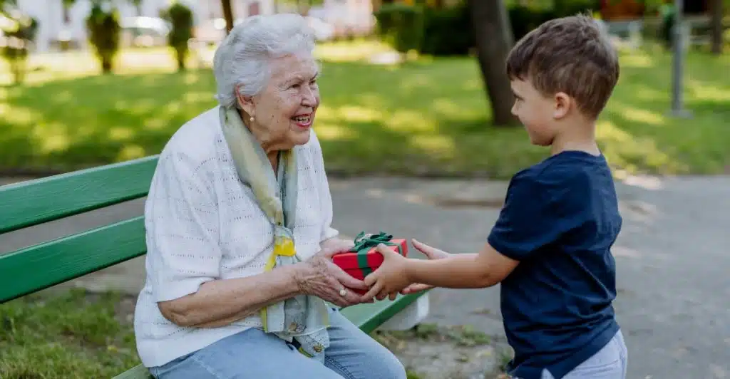 A young boy giving a wrapped gift to an older woman who is seated on a park bench.