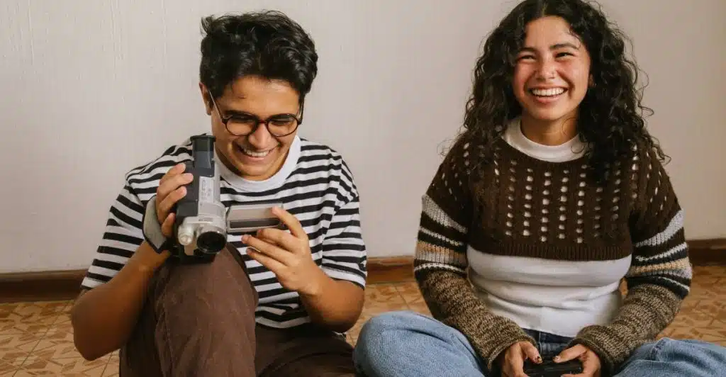  A young man with glasses holding a video camera and smiling with a young woman who is also smiling, sitting on the floor.
