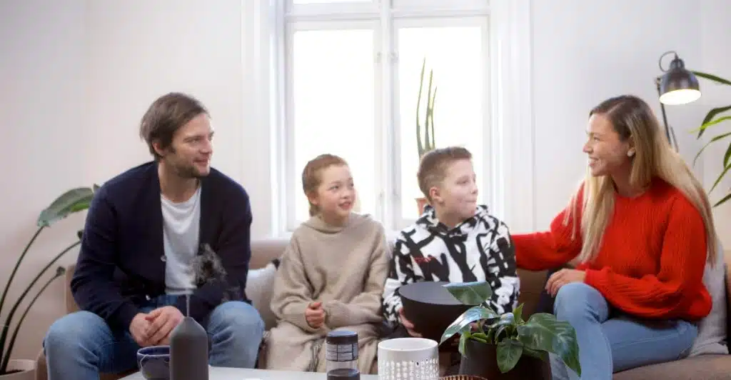A family of four, including parents and two young children, sitting together on a couch in a cozy living room.