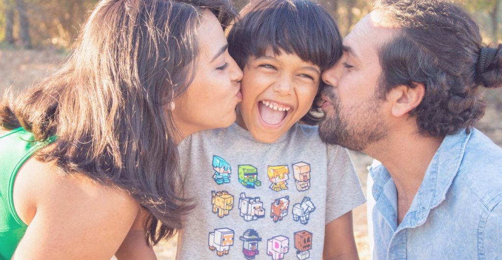 A mother and father kissing their smiling child on both cheeks outdoors.